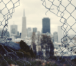 Looking through a hole in a chain link fence at a gray cityscape