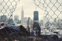 Looking through a hole in a chain link fence at a gray cityscape