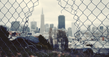 Looking through a hole in a chain link fence at a gray cityscape