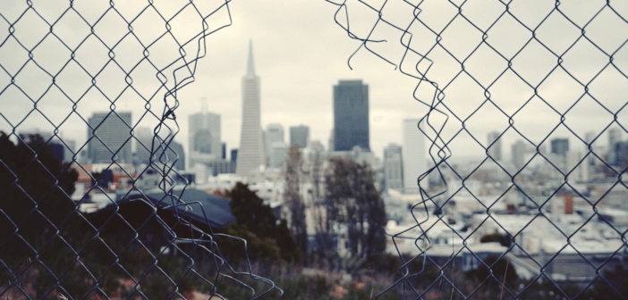 Looking through a hole in a chain link fence at a gray cityscape